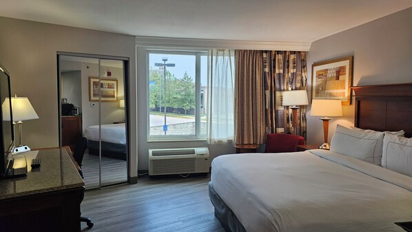 Una habitación de hotel con cama, escritorio con lámpara, silla, espejo, ventana con cortinas y vistas al exterior.