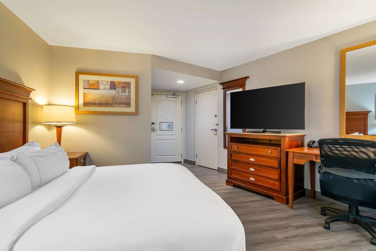 Ein Hotelzimmer mit einem großen Bett, einem Schreibtisch mit Stuhl, einem Fernseher und einer Tür zu einem anderen Raum.