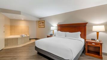 A hotel room with a large bed, a wooden headboard, two bedside tables with lamps, a bathroom with a bathtub, and a framed picture on the wall.