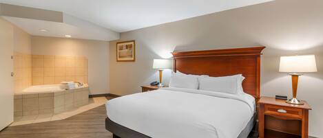 A hotel room with a large bed, a wooden headboard, two bedside tables with lamps, a bathroom with a bathtub, and a framed picture on the wall.