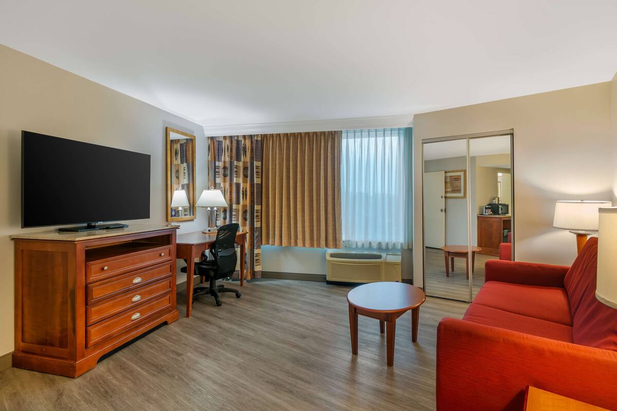 Ein Hotelzimmer mit einem Holzkoffer, einem Fernseher, einem Schreibtisch mit Stuhl, einem roten Sofa, einem kleinen Tisch und einem großen Spiegel.