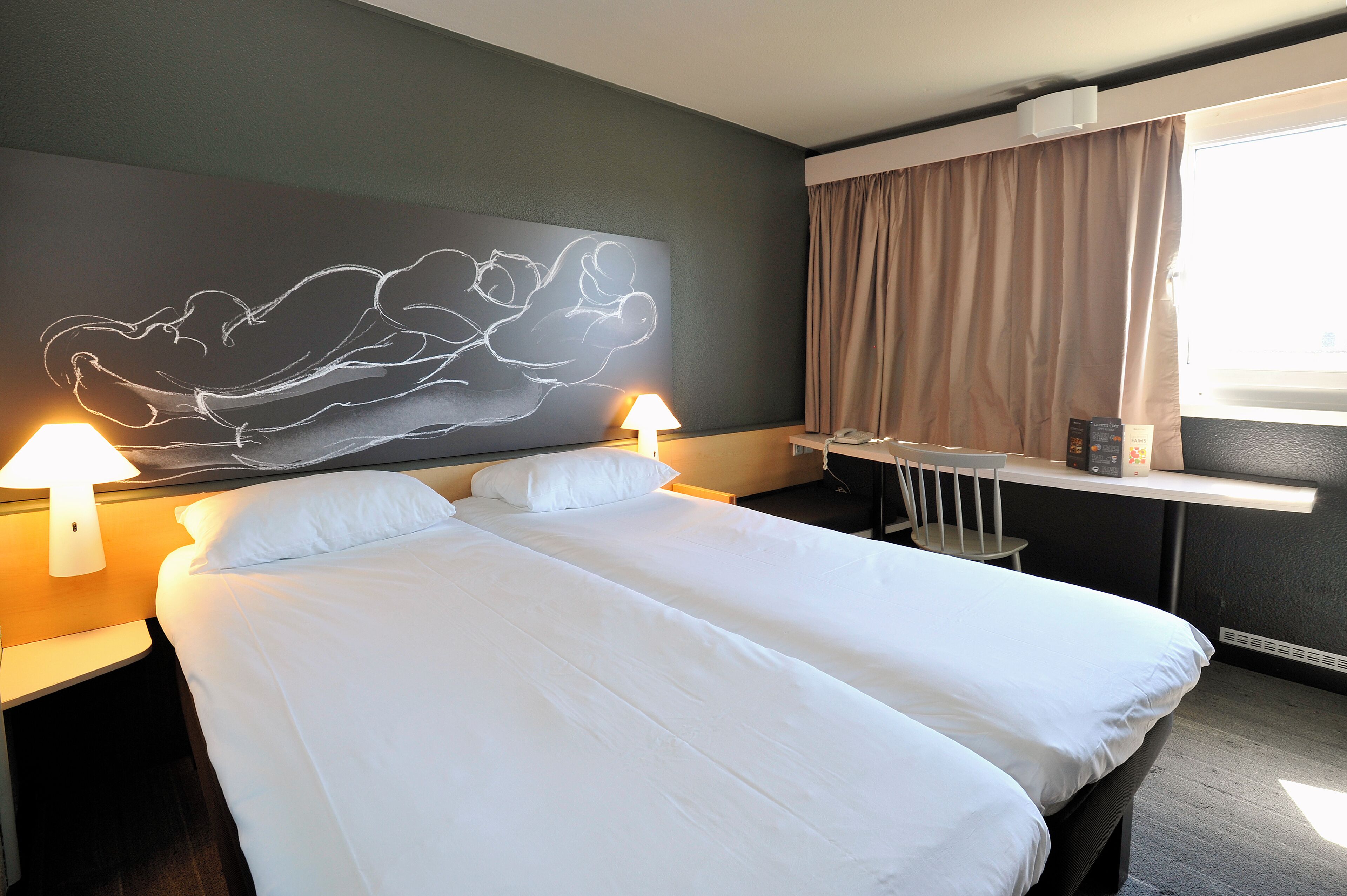 A hotel room with a large bed, two bedside tables with lamps, a desk with a chair, a window with curtains, and a wall art piece.