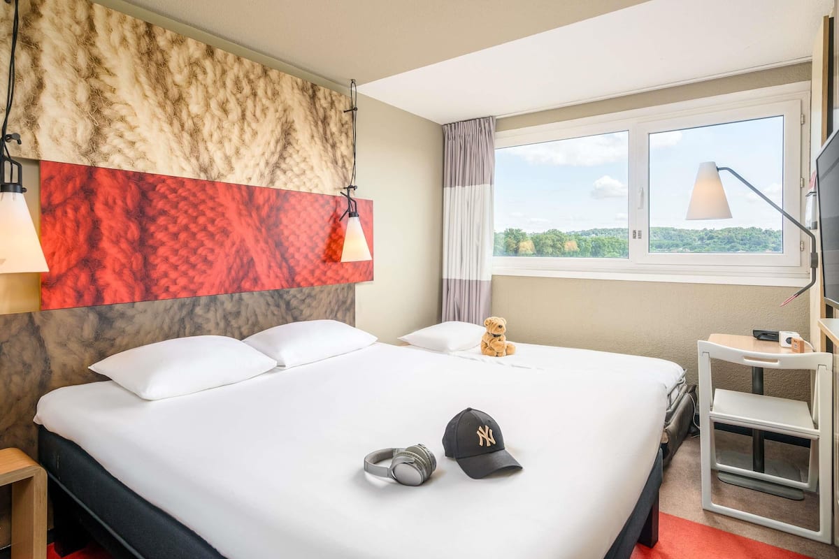 A hotel room with a large bed, a headboard with a textured design, a window with a view of greenery, a desk with a lamp, and a teddy bear on the bed.