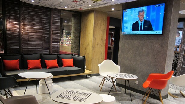 A modern lounge area with a sofa, chairs, and a TV screen displaying a news program. - ibis Lyon Villefranche sur Saone (Limas)