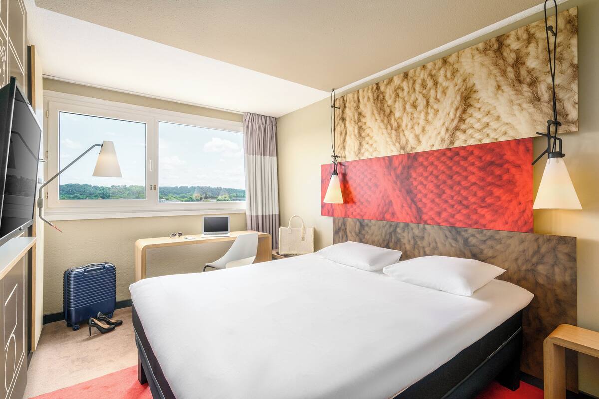 A hotel room with a bed, desk, chair, and a suitcase.