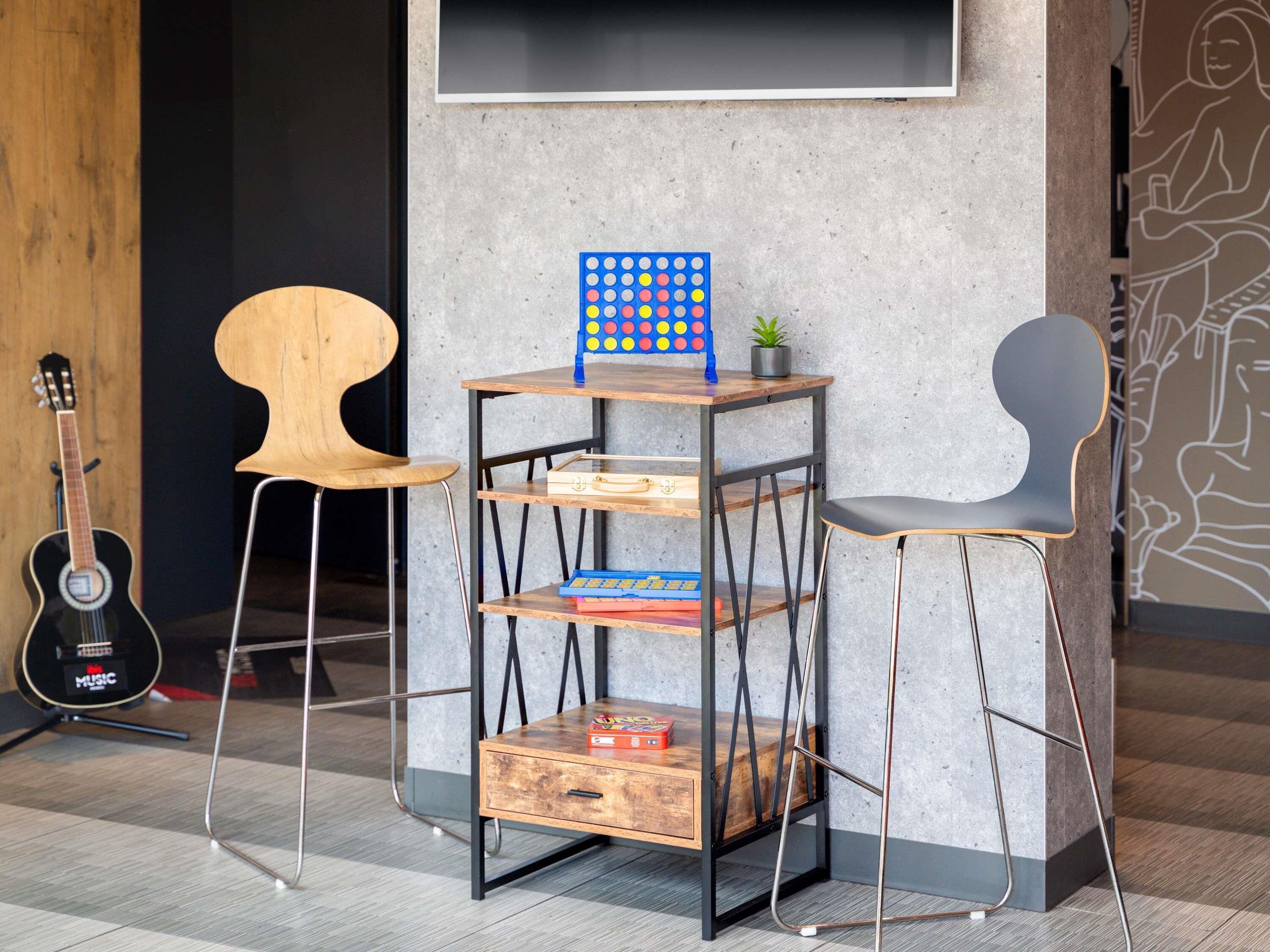A room with a wooden desk, a metal shelf unit, two chairs, a guitar, and a wall with a geometric pattern.