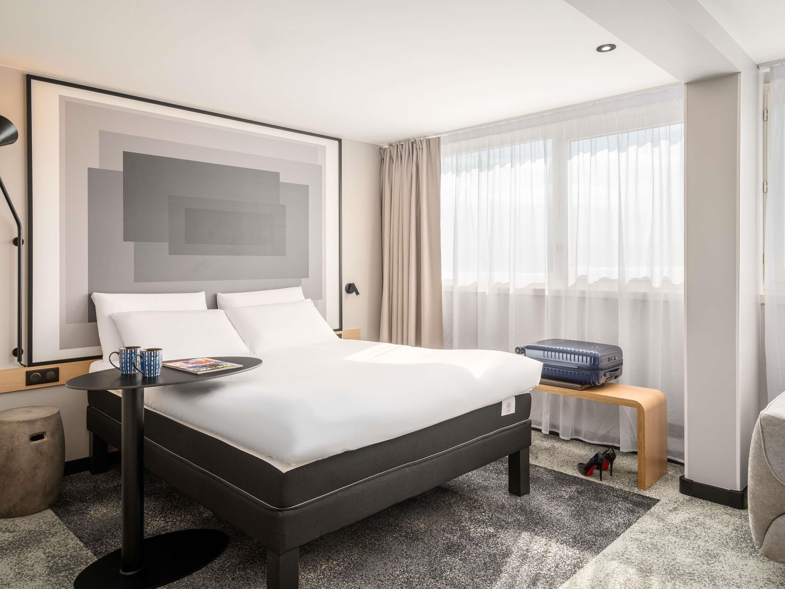 Una habitación de hotel moderna con una cama grande, una mesita de noche, una lámpara y una ventana con cortinas.