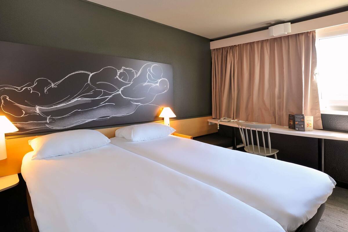 A hotel room with a bed, bedside lamps, a desk with a phone, and a window with curtains.