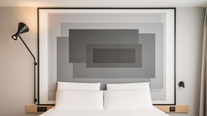 A neatly made bed with white linens, a wall-mounted picture frame with geometric design, a black floor lamp, and a wooden bedside table. - ibis Lyon Villefranche sur Saone (Limas)