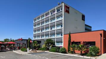 A modern hotel with a multi-story structure, a red-brick building adjacent to it, and a parking lot with red umbrellas.