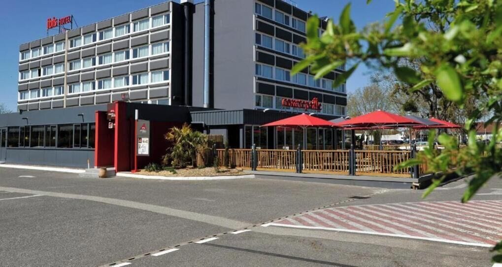 Ein Hotel mit moderner Fassade, einem Parkplatz und Sitzgelegenheiten im Freien mit roten Sonnenschirmen.