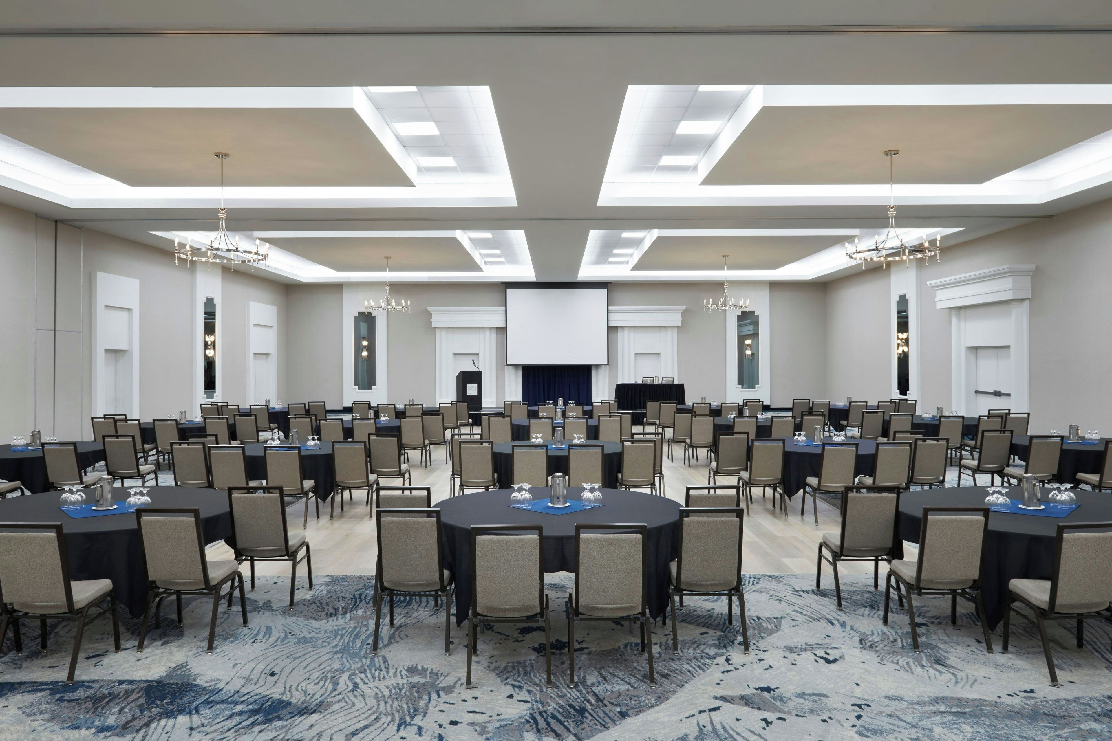A large conference room with multiple tables and chairs, a central screen, and chandeliers.
