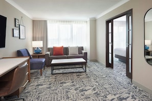 A hotel room with a sofa, a desk, a chair, a coffee table, a bed, a large window with curtains, and a door leading to another room. - Four Points by Sheraton London (London)