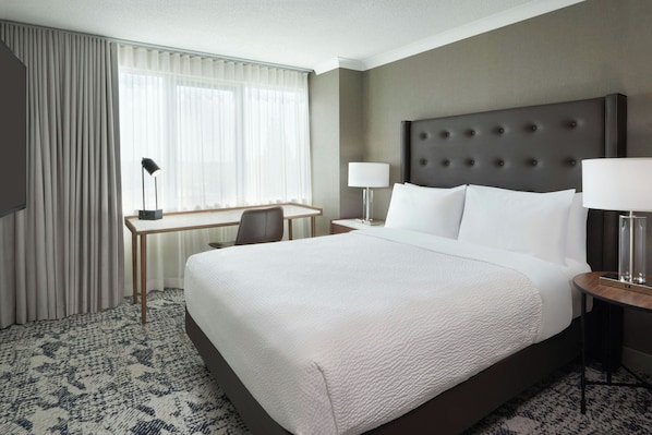A hotel room with a large bed, a desk, a chair, two bedside lamps, and a window with curtains.