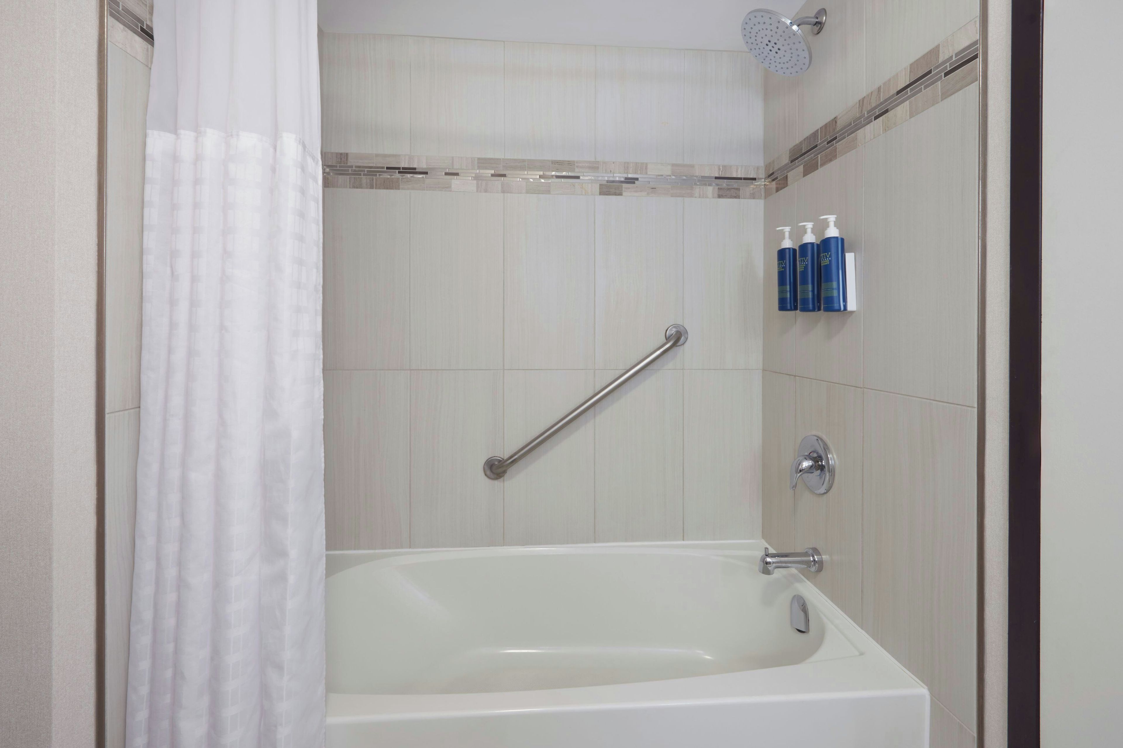 A bathroom with a bathtub, a showerhead, a handrail, and toiletries on the wall.