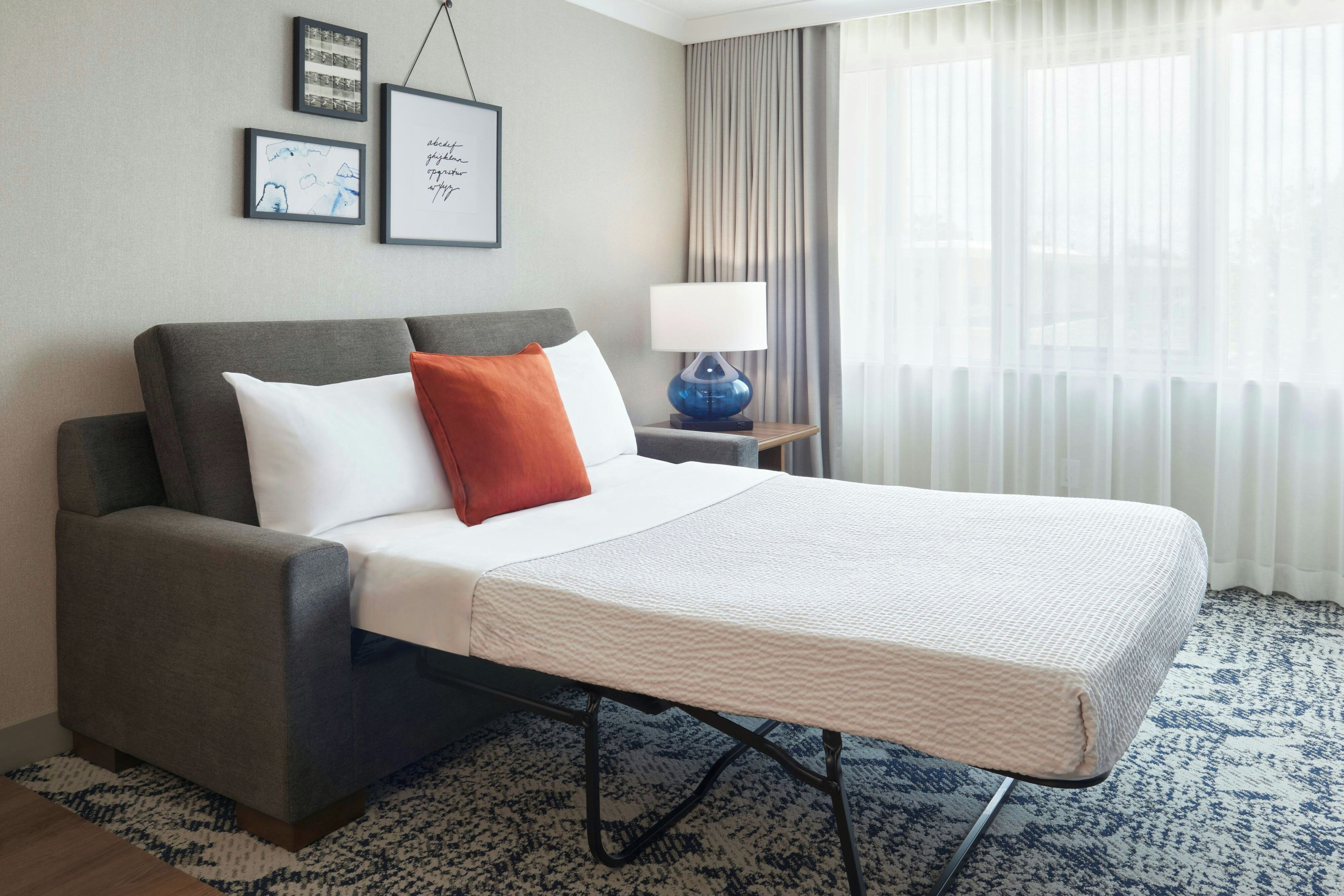 A hotel room with a bed, a nightstand, a lamp, a window with curtains, and framed pictures on the wall.