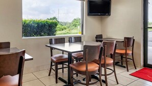 A dining area with tables and chairs, a television mounted on the wall, and a window with a view of greenery. - Econo Lodge Sebring (Avon Park)