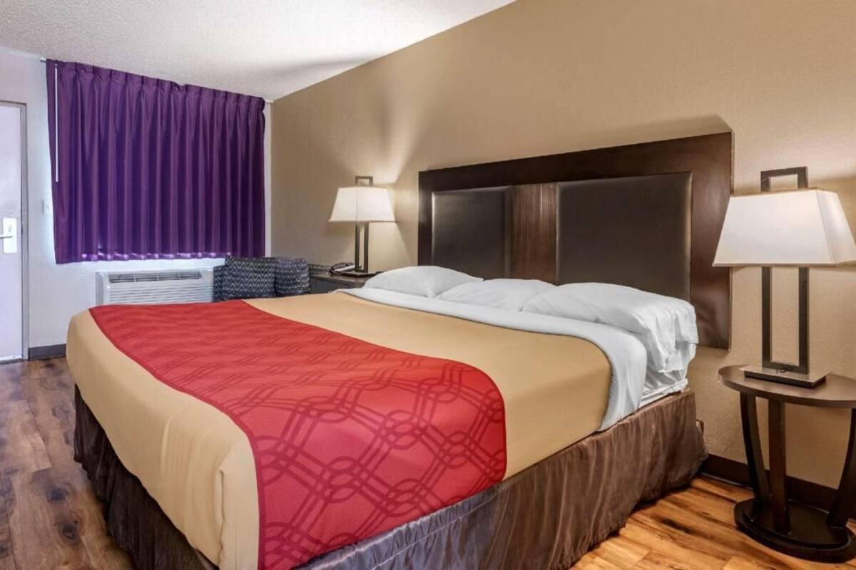 A hotel room with a bed, two bedside lamps, a window with purple curtains, and an air conditioning unit.