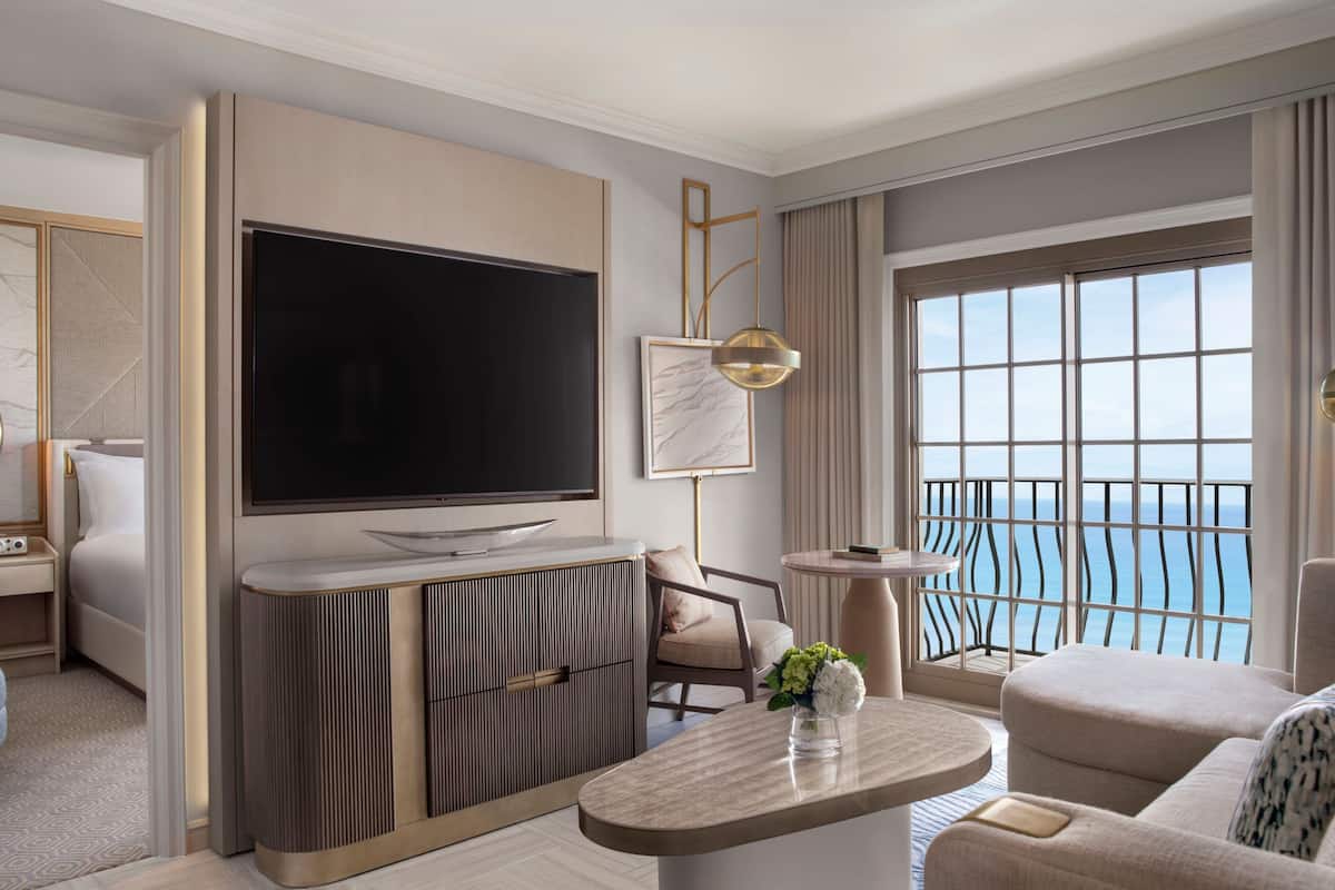 Ein modernes Hotelzimmer mit einem großen Flachbildfernseher, einer Couch, einem kleinen runden Tisch und einem Balkon mit Meerblick.