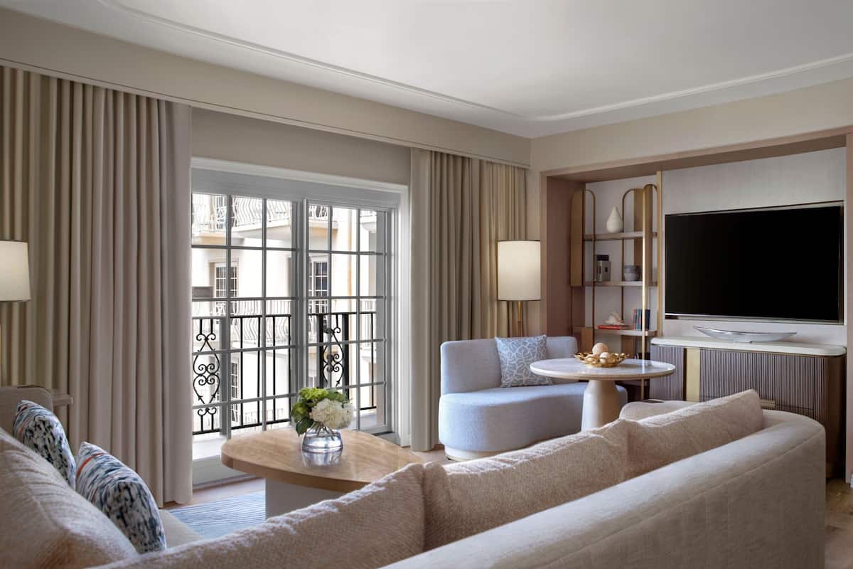 Ein modernes Wohnzimmer mit Sofa, Sessel und Flachbildfernseher. Ein Balkon mit Geländer und Vorhängen ist vorhanden, sowie ein kleiner Tisch mit Blumenvase.