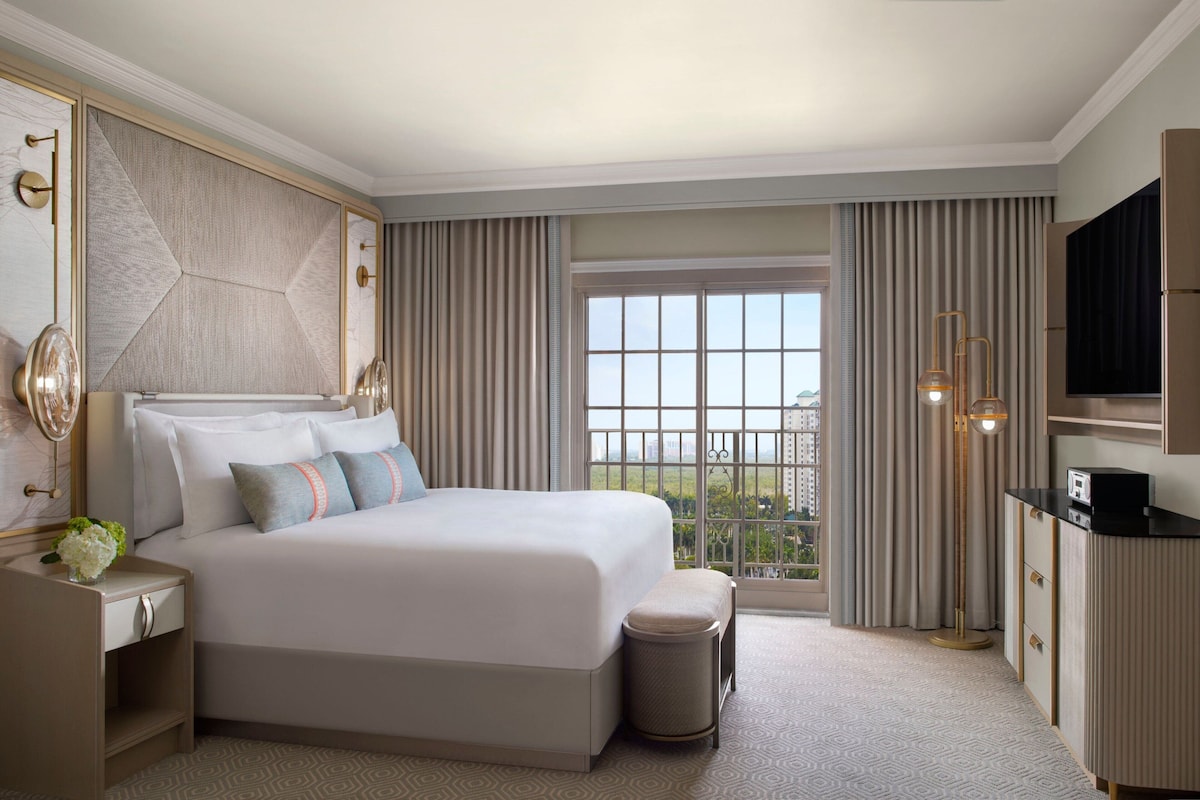 A hotel room with a large bed, a desk, a TV, and a view of the city.