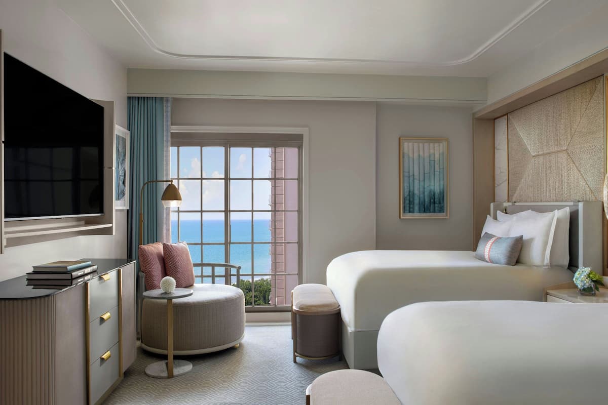 Ein Hotelzimmer mit zwei Betten, einem Schreibtisch, einem Sessel, einem Fernseher und Meerblick.