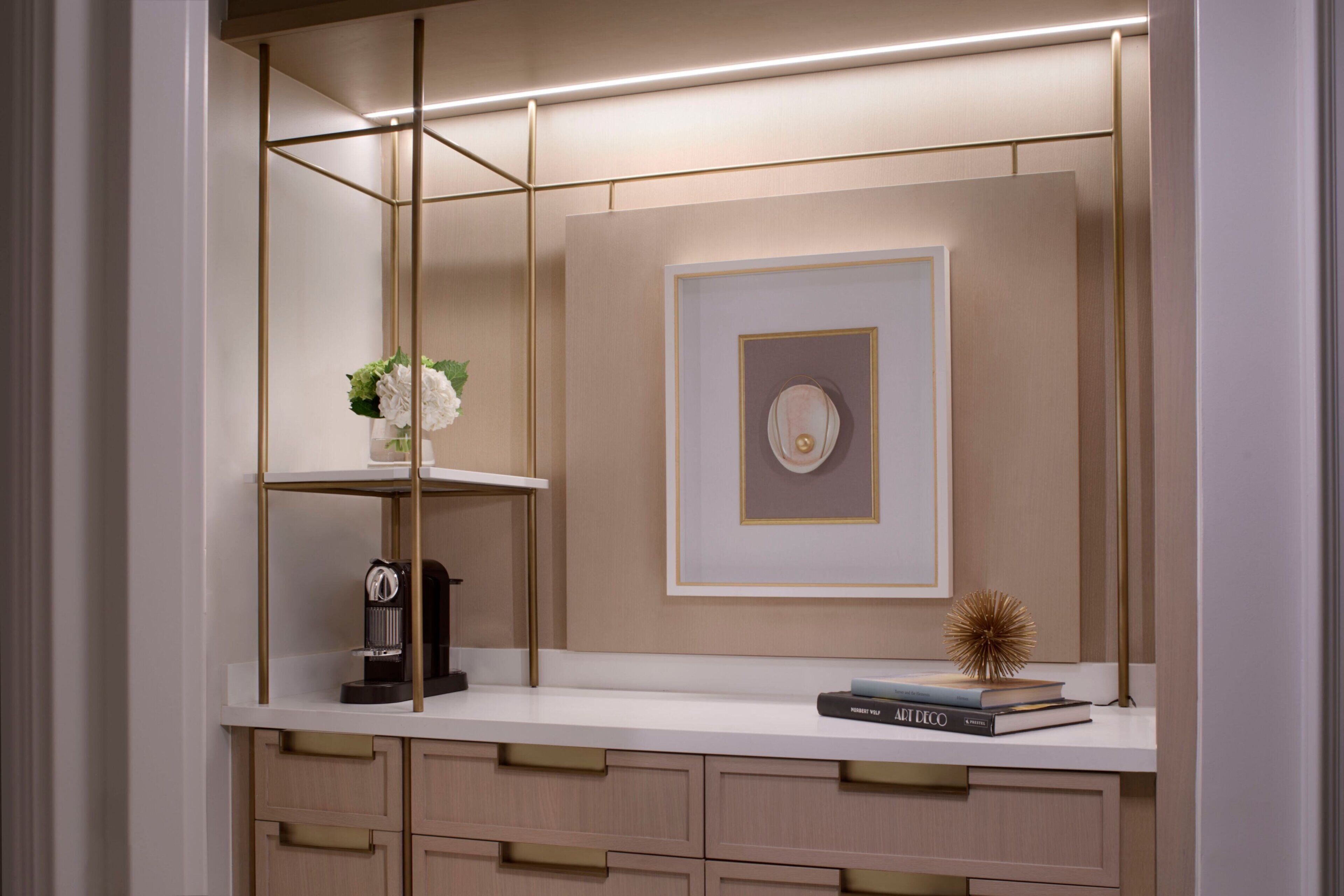 A modern, well-lit room with a white countertop, a coffee machine, a vase with flowers, a framed picture, and a book titled 'ART DECO'.