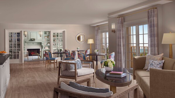 A spacious living room with a fireplace, a dining area with floral chairs, and a view of the sea through large windows.
