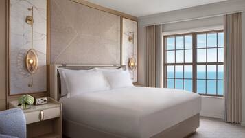 A large bed with a headboard, two bedside tables with clocks, and a view of the sea through a large window.
