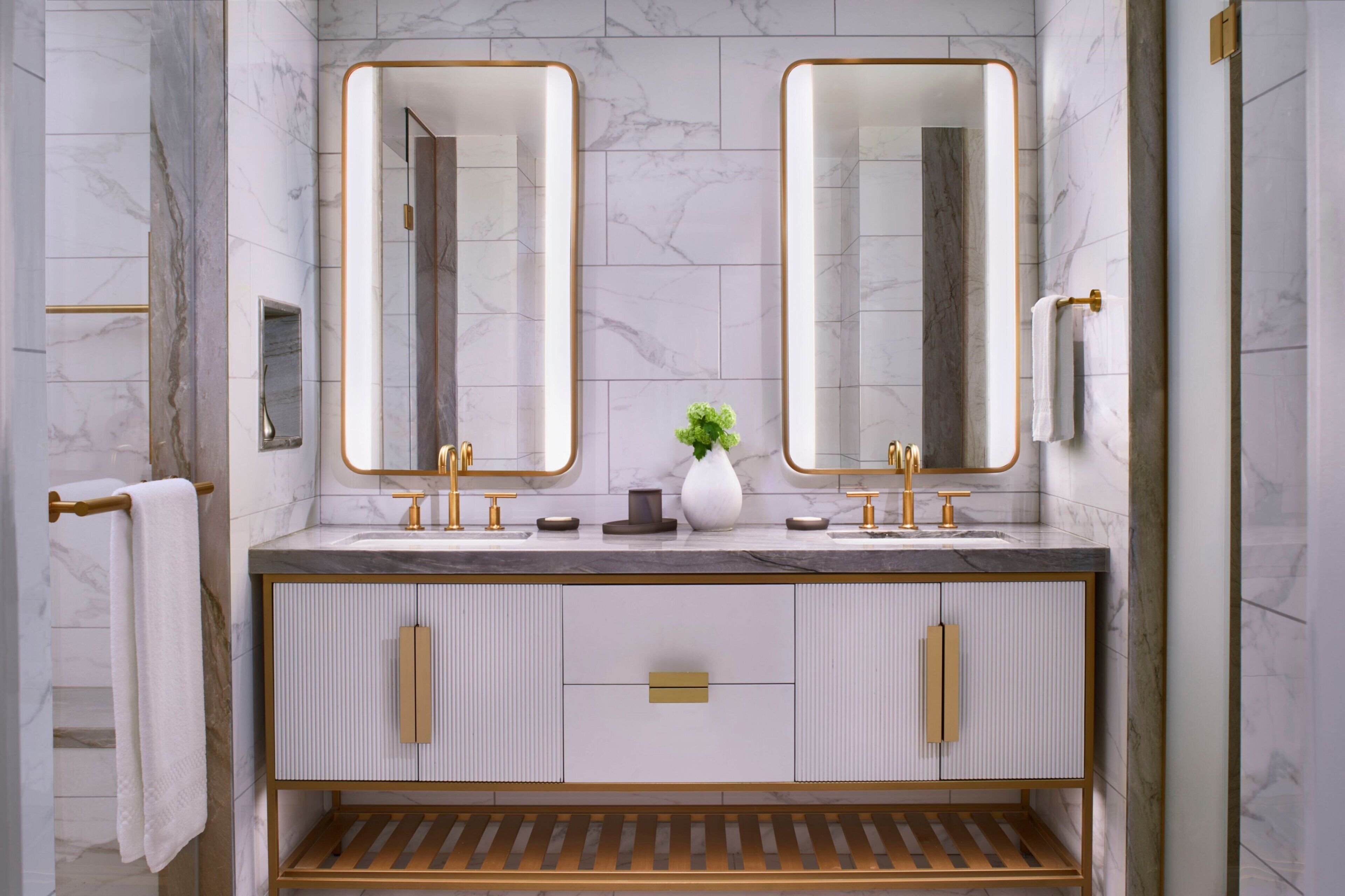 A modern bathroom with two large mirrors, a white vanity with gold handles, and a wooden shelf.