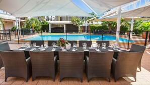 An outdoor dining area with tables set for six, surrounded by wicker chairs, under a white canopy, with a pool in the background. - ibis Styles Karratha (Karratha)