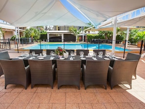 Un espace de restauration en plein air avec des tables disposées pour accueillir six personnes, entouré de chaises en osier, sous une tonnelle blanche, avec une piscine en arrière-plan.