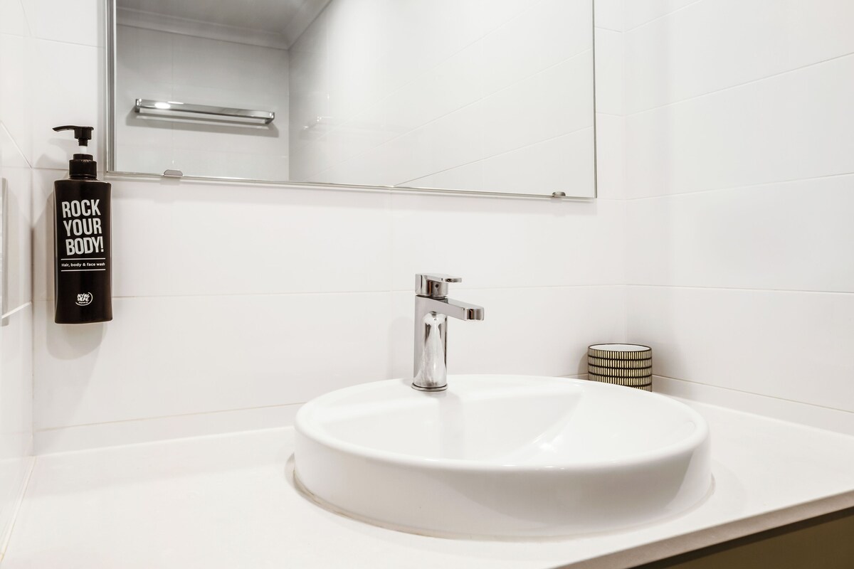 A bathroom with a white sink, a mirror, a faucet, and a bottle of body wash.