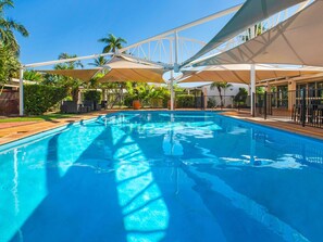 A swimming pool with a clear blue water surface, surrounded by palm trees and shaded by large umbrellas. - ibis Styles Karratha (Karratha)