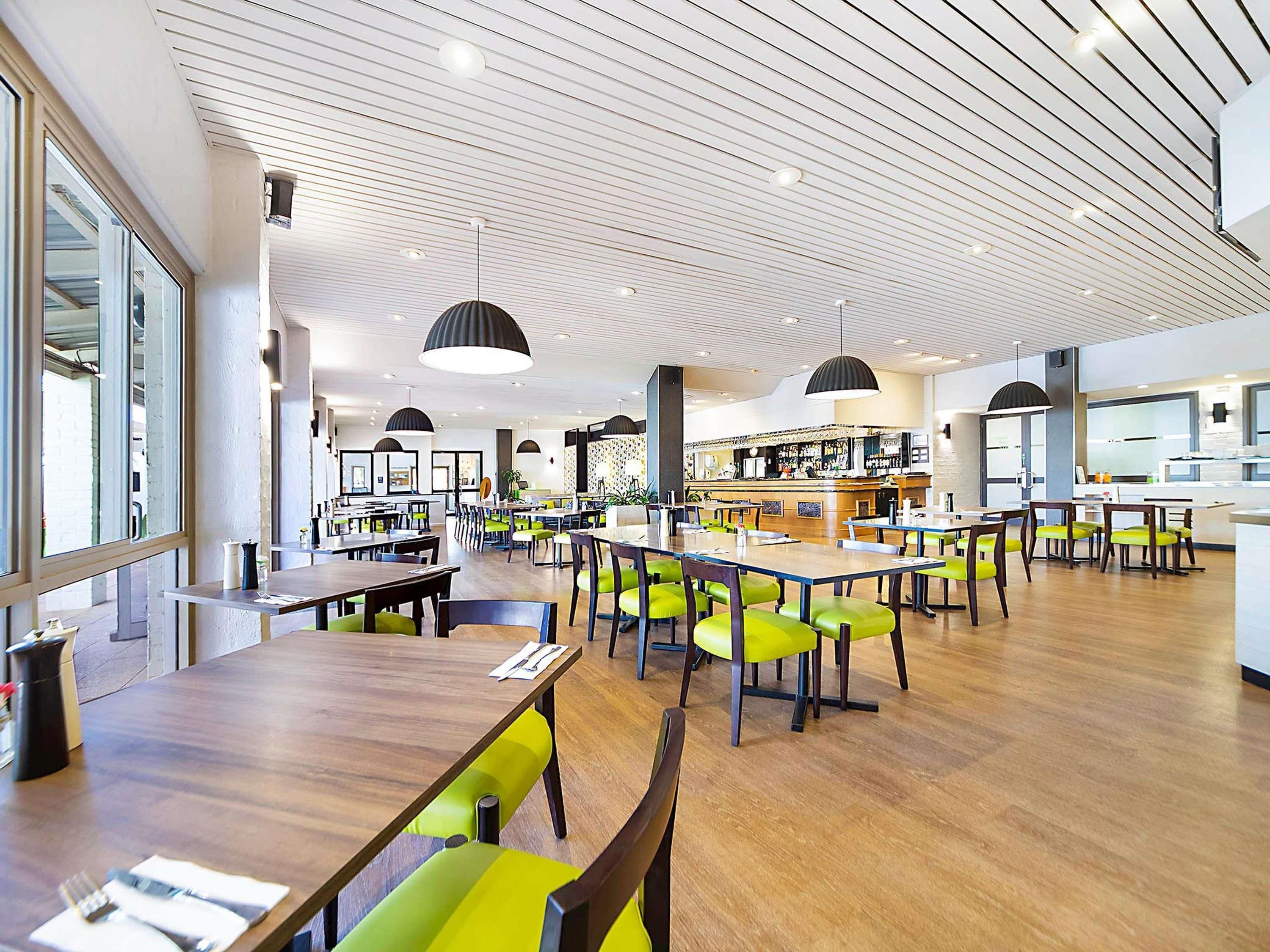 Un comedor amplio con mesas de madera y sillas verdes, una barra al fondo y un diseño de techo moderno.
