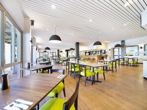 Ett rymligt matområde med träbord och gröna stolar, en bar i bakgrunden och en modern takdesign.