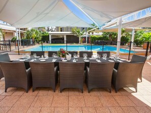 An outdoor dining area with tables set for six, surrounded by wicker chairs, under a white canopy, with a pool in the background. - ibis Styles Karratha (Karratha)