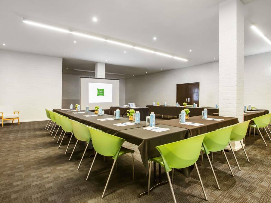 Uma sala de conferências com uma mesa comprida, cadeiras verdes e uma tela de projetor exibindo 'ibis Styles'.