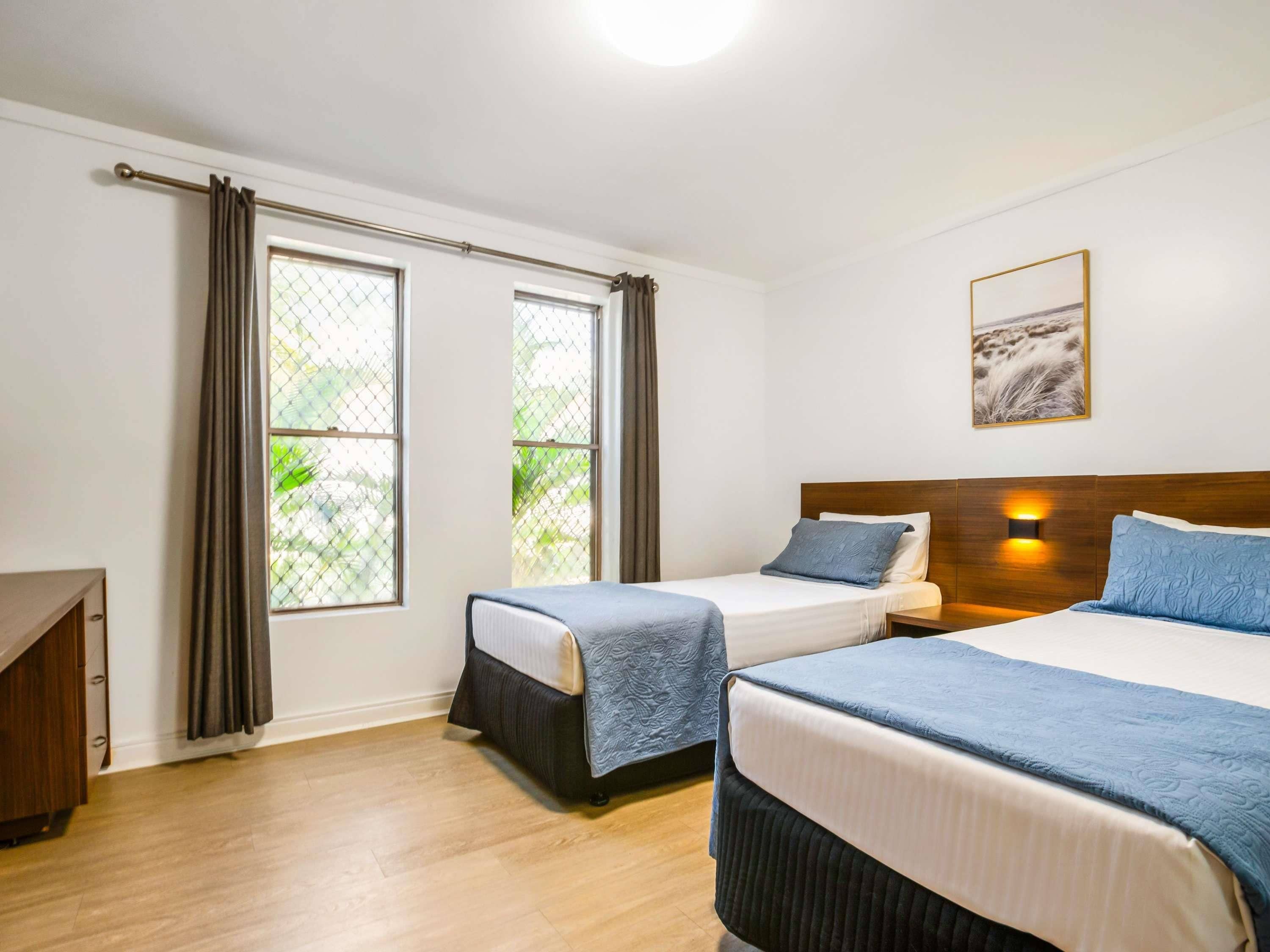 A hotel room with two beds, a wooden headboard, a nightstand, a desk, and a window with curtains.