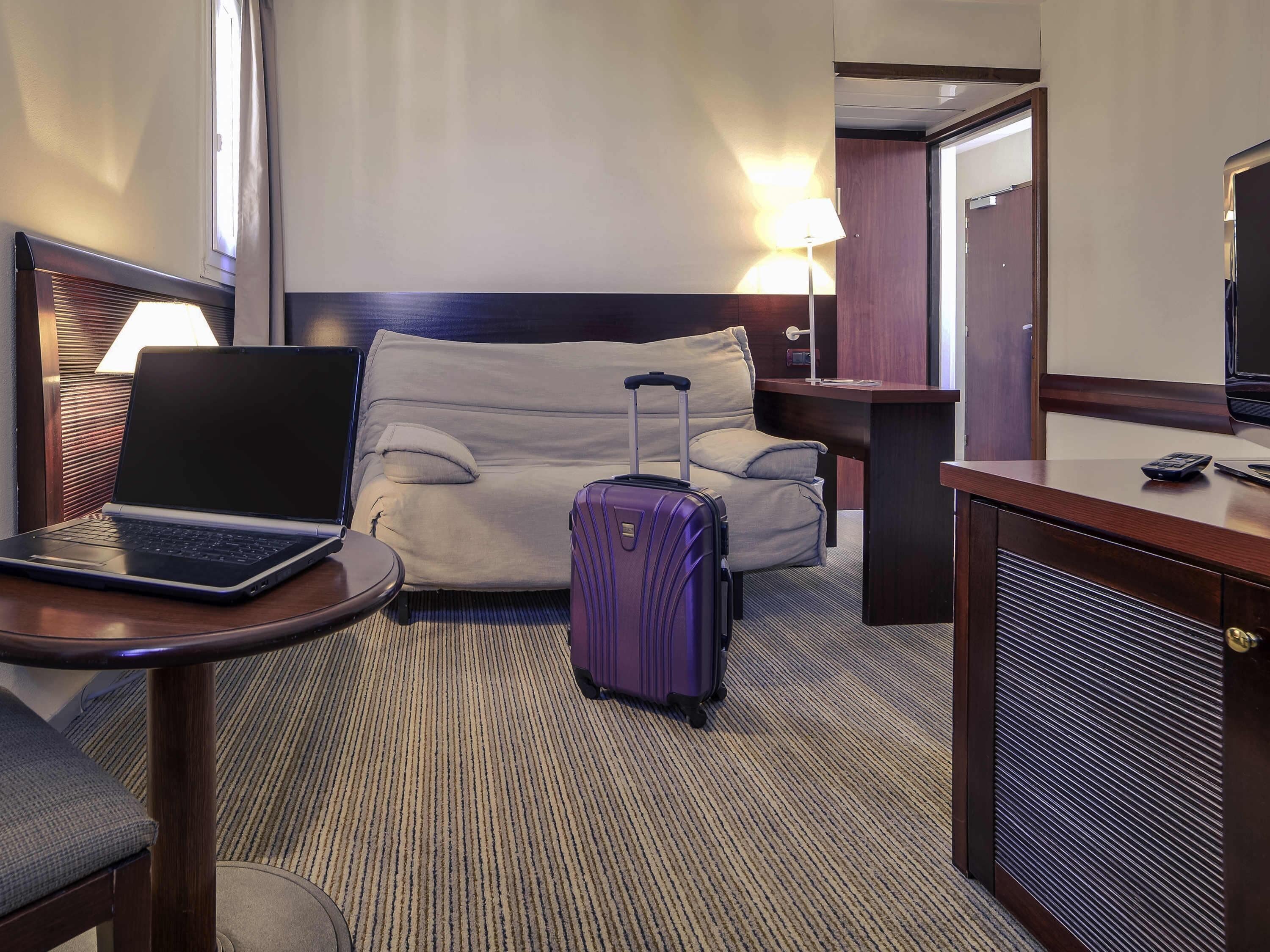 A hotel room with a bed, a desk with a laptop, a purple suitcase, a lamp, and a television.