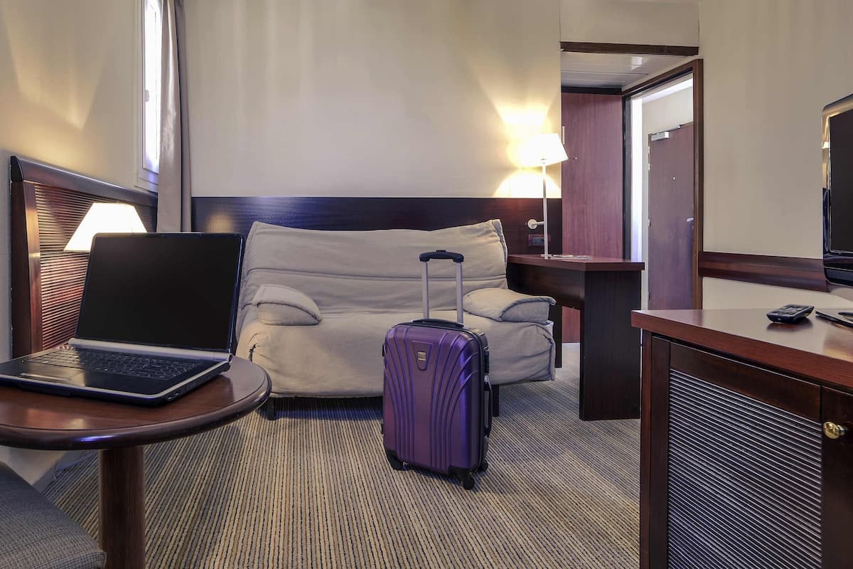 A hotel room with a bed, a desk with a laptop, a purple suitcase, a lamp, and a television.