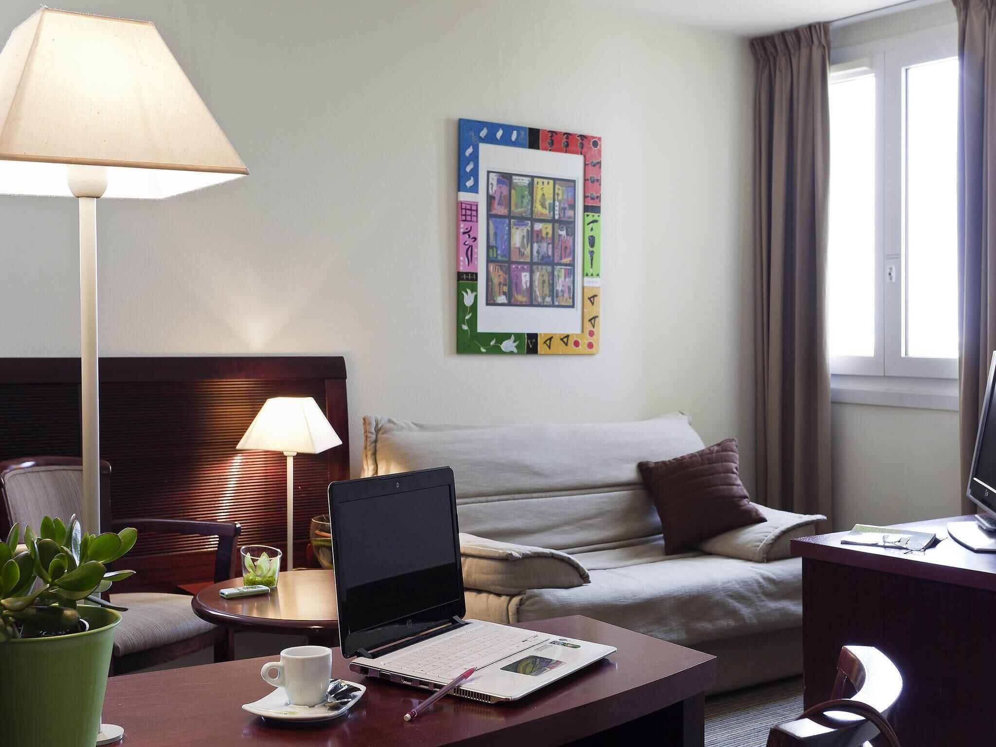 Une chambre d’hôtel avec un lit, un bureau avec un ordinateur portable, une lampe, une plante et une œuvre d’art murale.