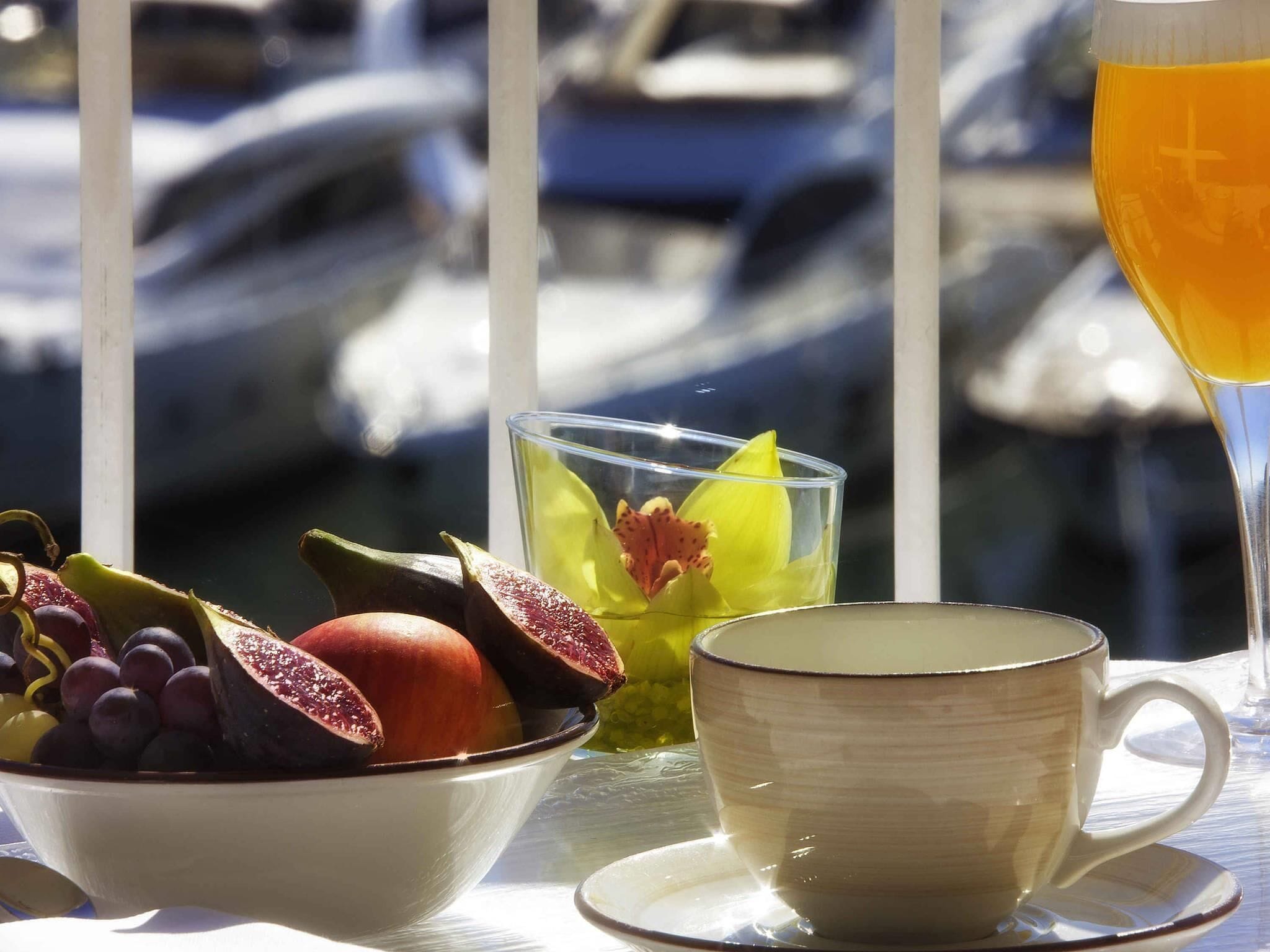 Ein Tisch mit einer Obstschale, einem Glas Orangensaft und einer Tasse, vor einer Ansicht von geparkten Autos.
