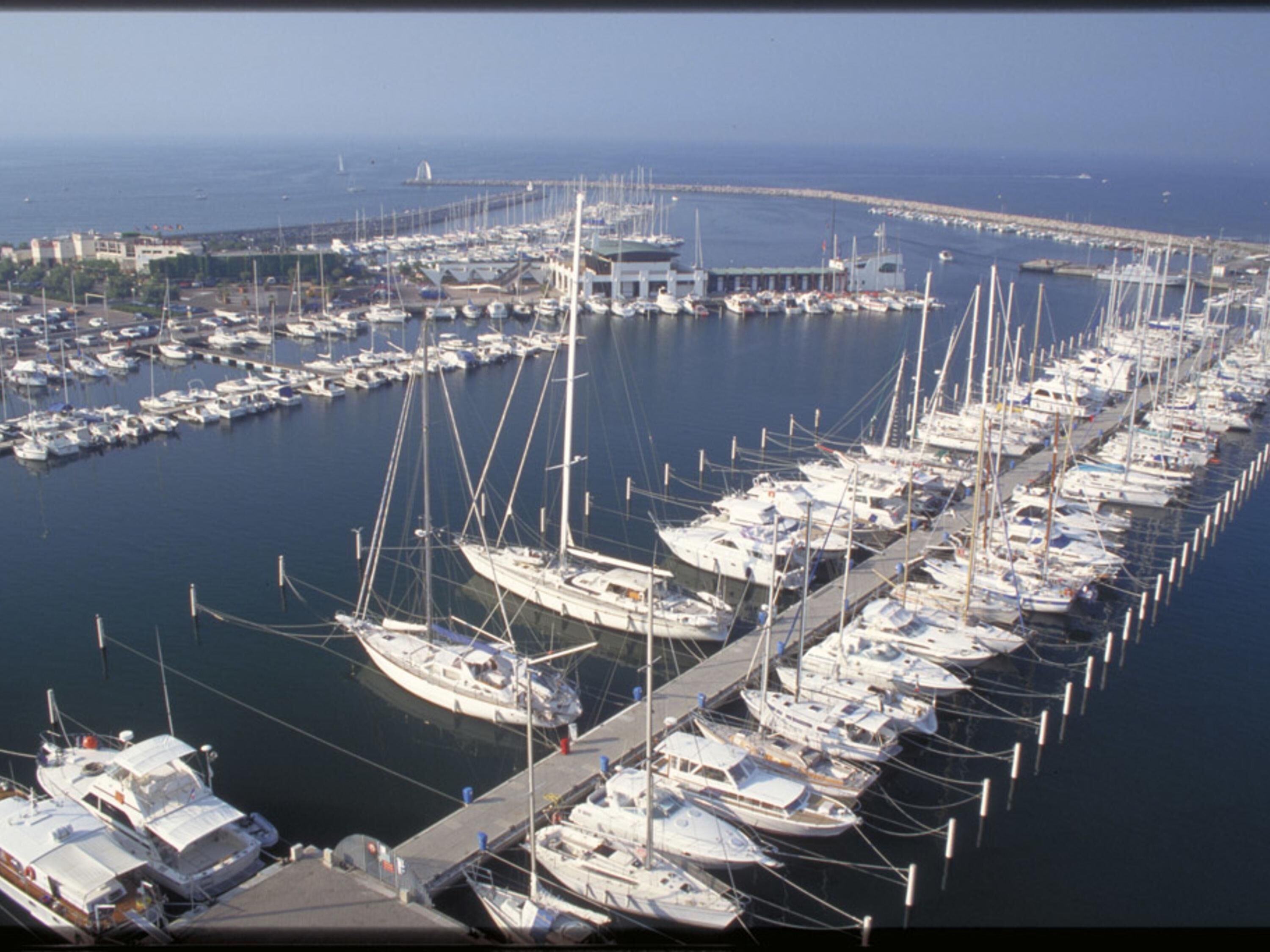 A marina with numerous boats docked in a row, a pier extending into the water, and a distant view of the coastline.