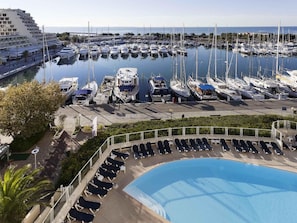 A marina with numerous boats docked, a swimming pool with sun loungers, and a large building with balconies.