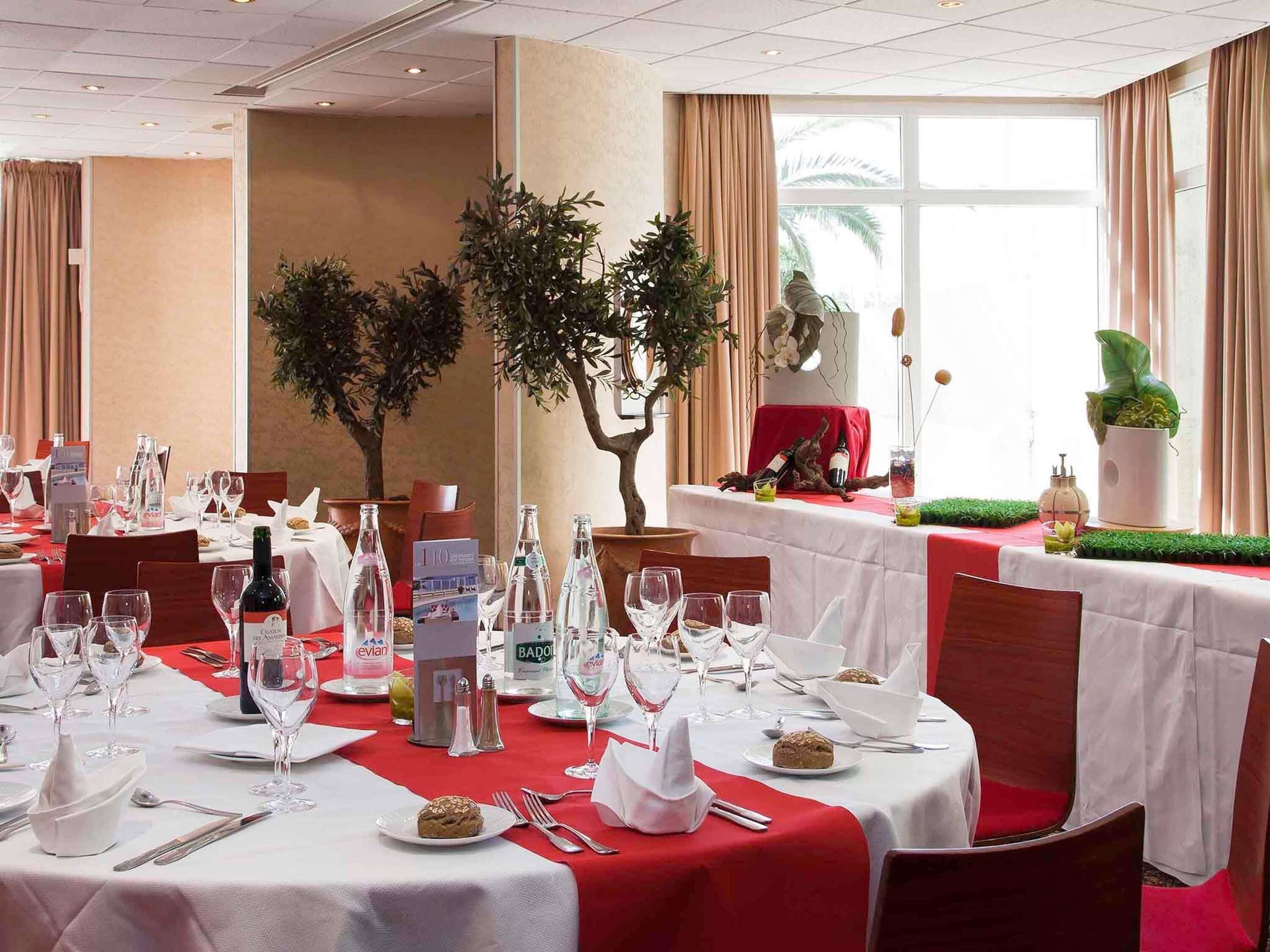 A hotel dining area with round tables set for a meal, featuring white tablecloths, red accents, and a variety of tableware.