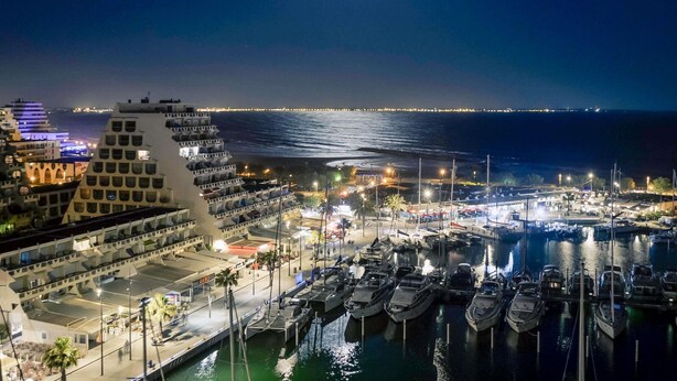A marina with boats docked, a waterfront promenade, and a cityscape with illuminated buildings in the background. - Mercure Grande Motte Port (La Grande-Motte)