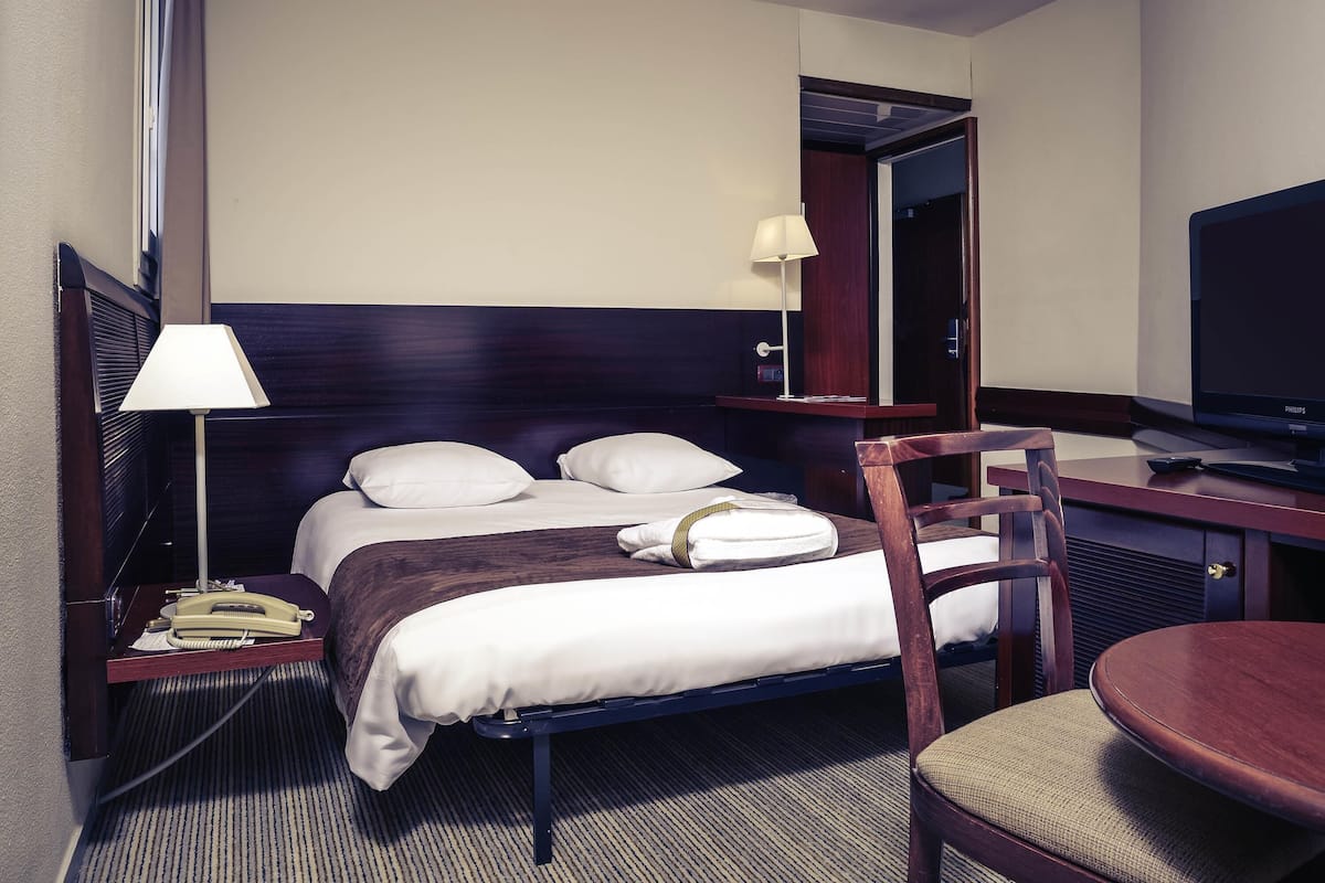 A hotel room with a bed, bedside table with a lamp, a chair, a desk with a television, and a mirror.