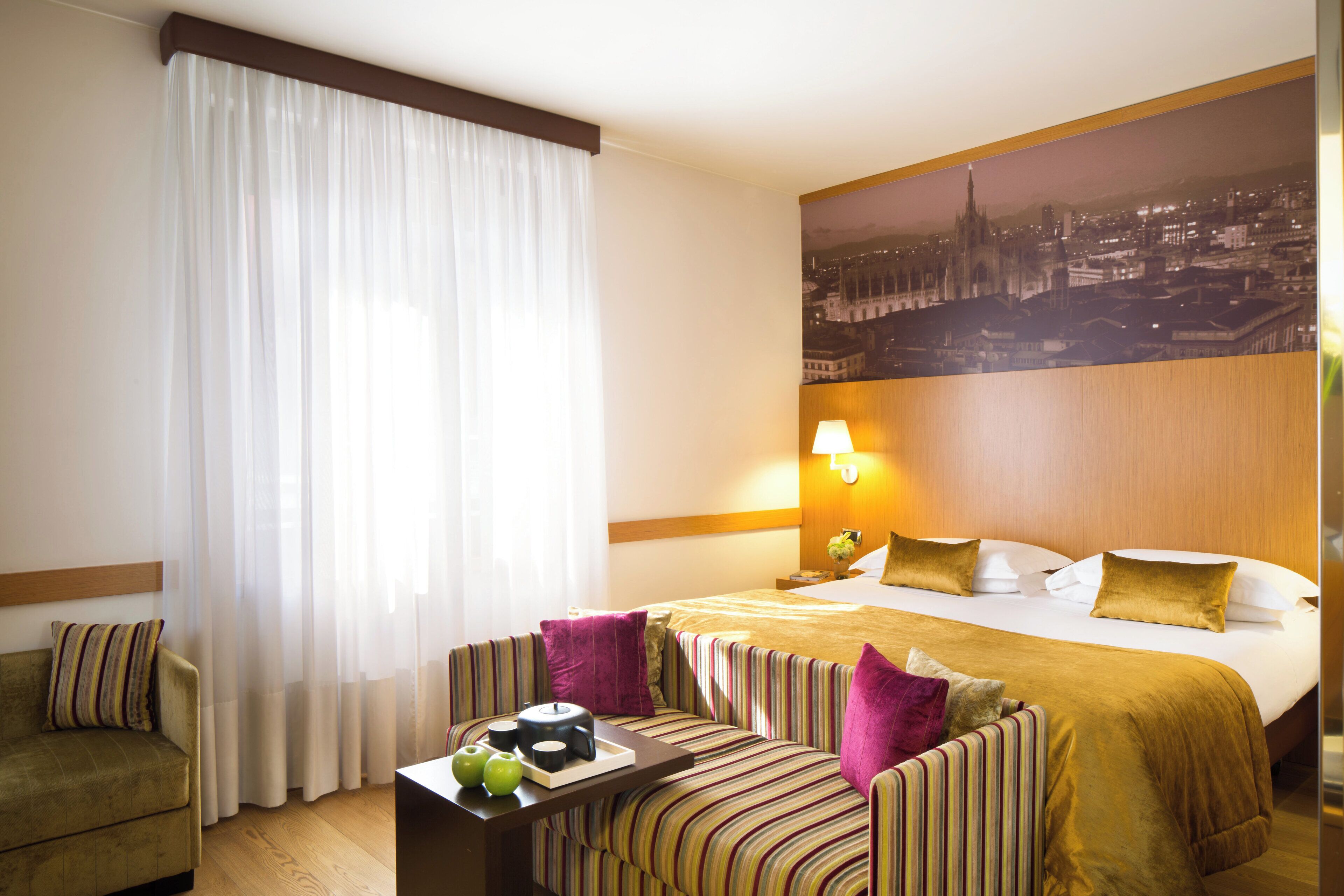 Quarto de hotel com uma cama, um sofá, uma mesinha com maçãs e uma pintura da paisagem urbana na parede.