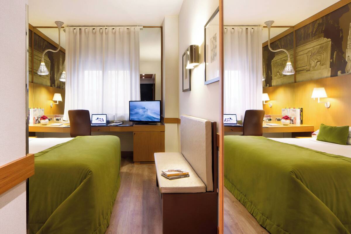 A hotel room with two single beds, a desk with a computer, a chair, and a small table.